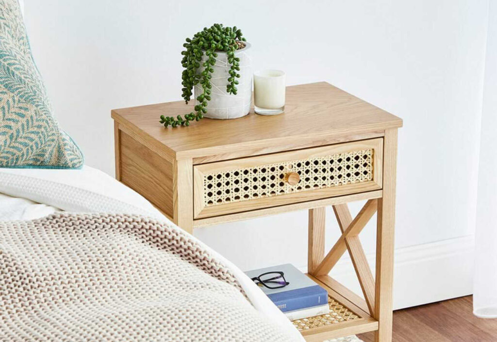 Hamptons-style wooden bedside table with a plant on the top next to a bed.