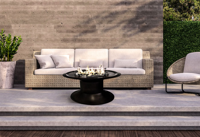 Black gas firepit sits on a concrete patio next to an outdoor sofa.