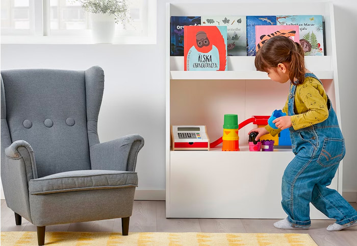 Little girl in denim overalls plays with toys on a white bookcase.