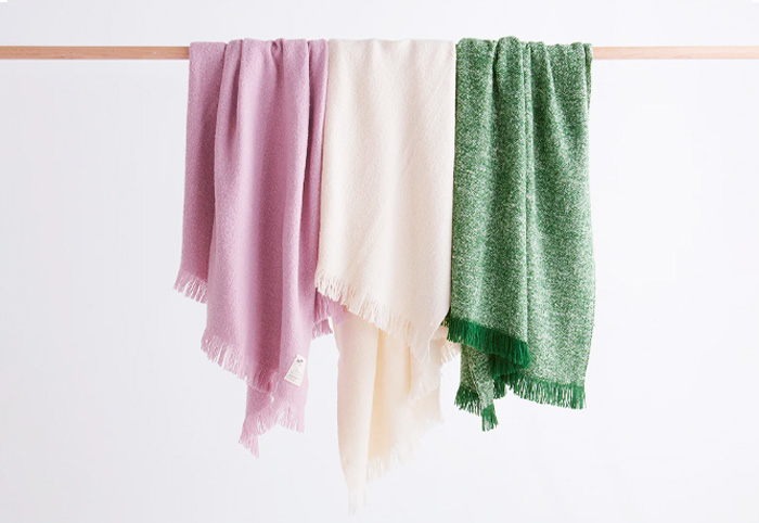 Mauve, natural and green wool blankets hanging from a wooden pole.