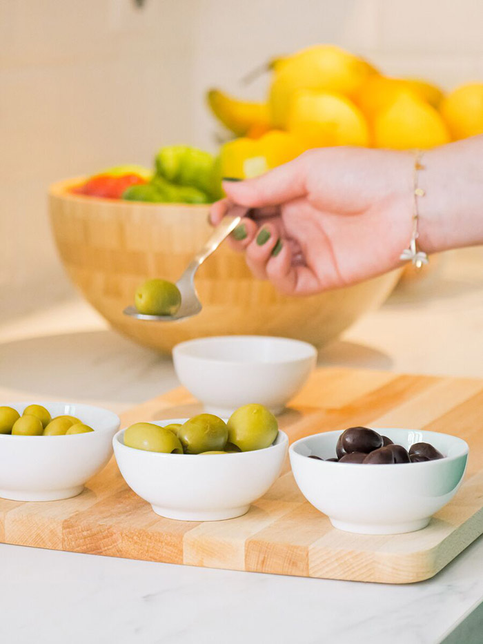 Woman spoons olives from small serving bowls on a charcuterie board.