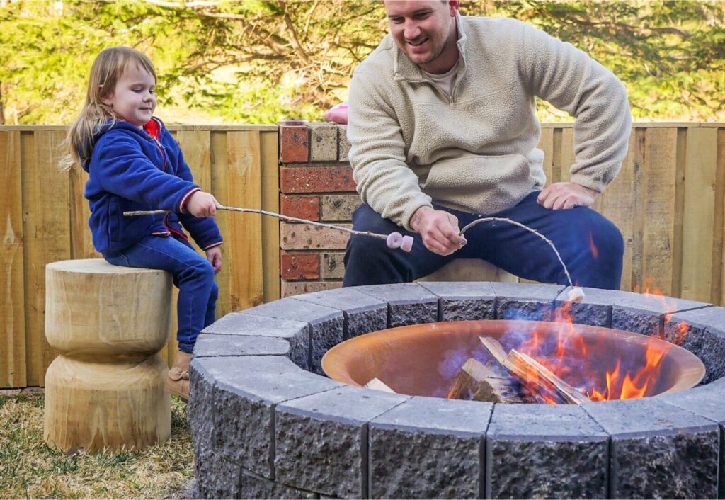 Father and child smile as they toast marshmallows at a fire pit.