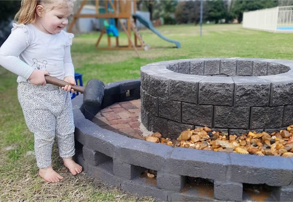 Small child holds a hammer next to a half-built fire pit.