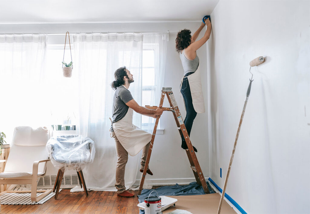 Man holding ladder as woman tapes a ceiling ready for painting.