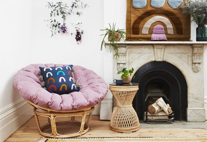 Rattan occasional chair with a fluffy pink cushion in a living room next to an ornate fireplace.