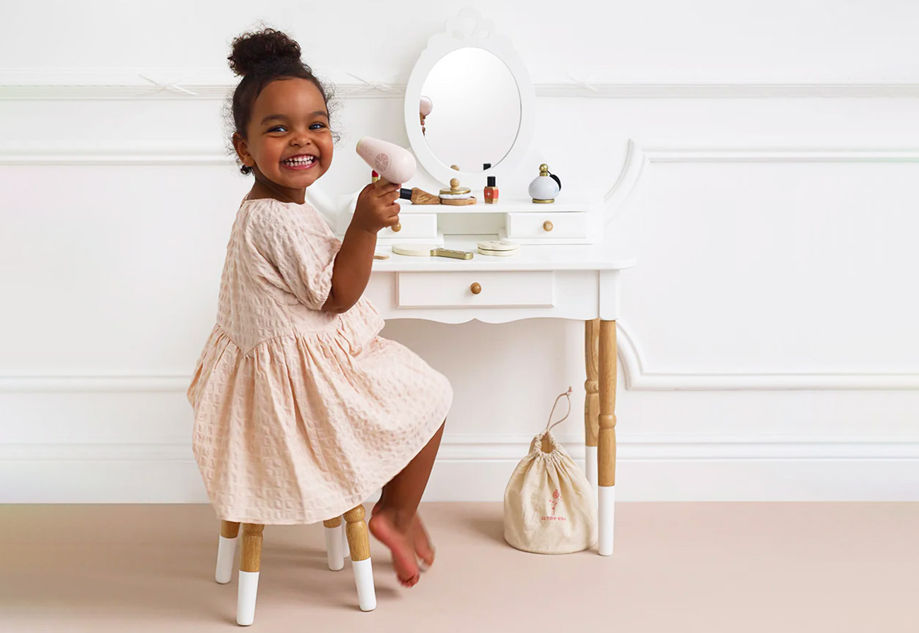 Smiling child sitting at a white kids' dressing table holding a toy hairdryer.