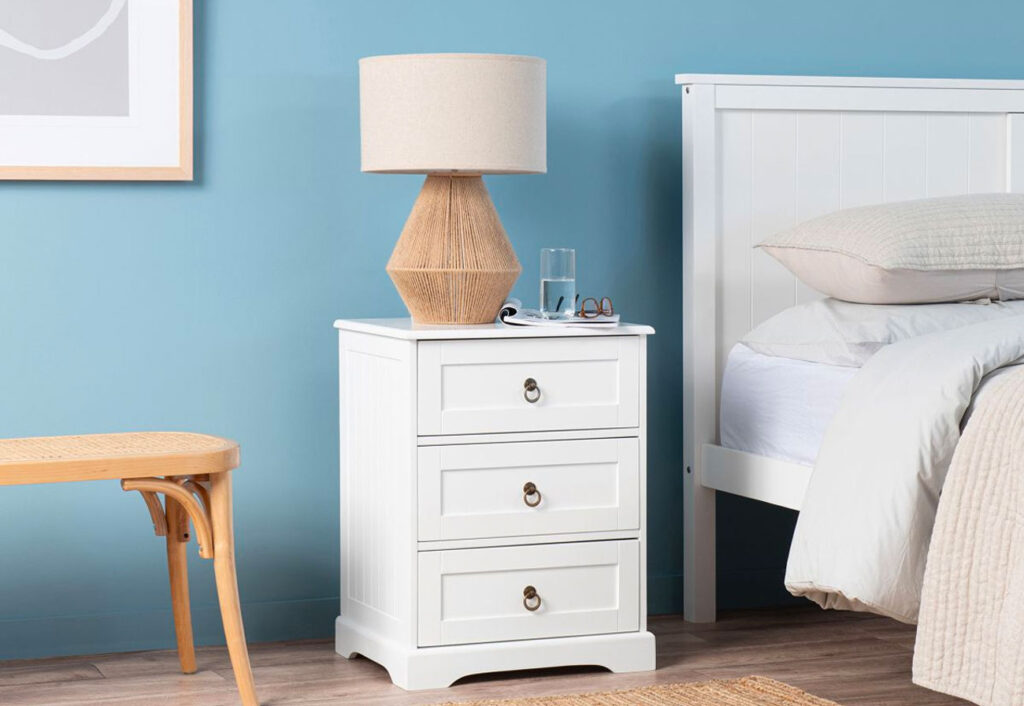 White bedside table sits against a blue wall with a rattan lamp on top of it.