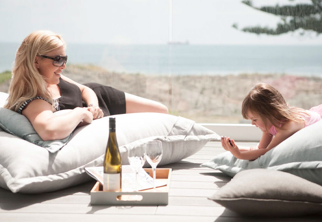 A woman and a child lounge on cushions on a deck overlooking the beach.
