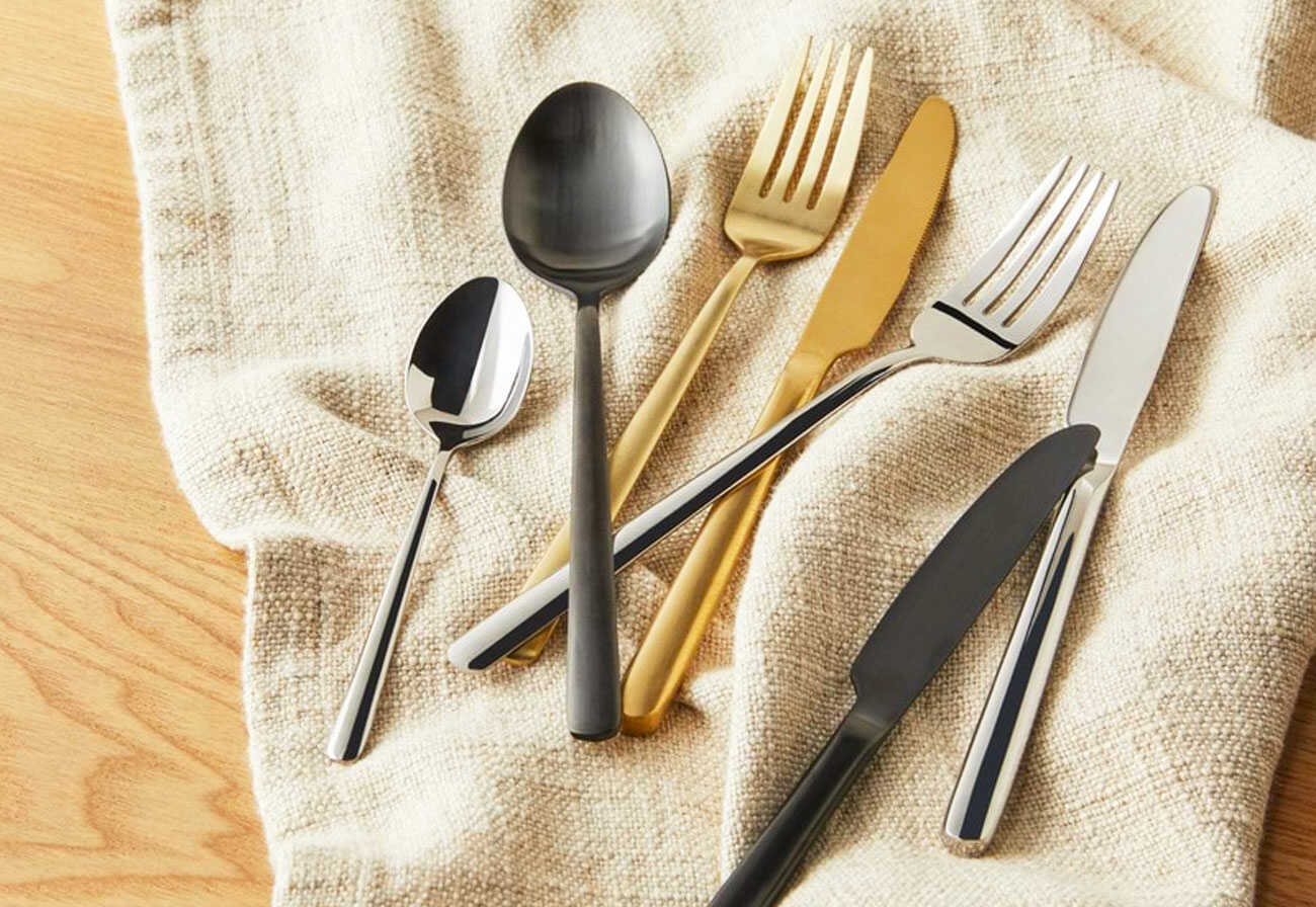 Black, gold and silver cutlery on top of a cream tea towel on a wooden table.