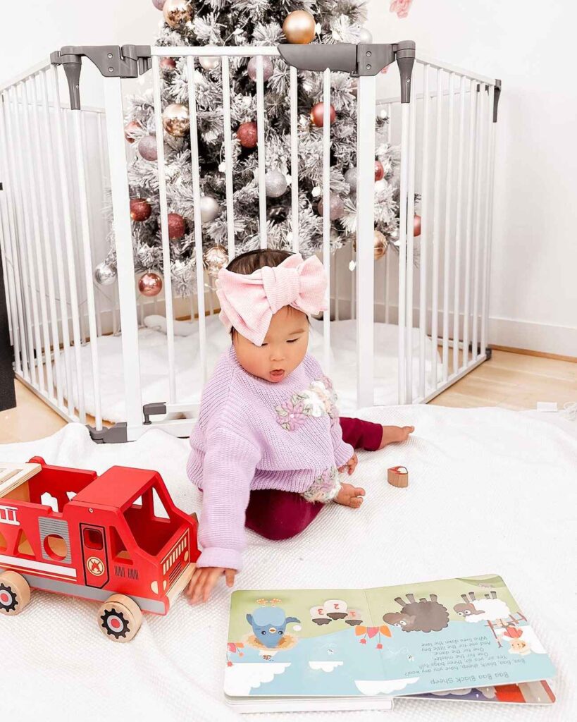 Baby playing on a white mat in front of a christmas tree that's enclosed in a playpen.