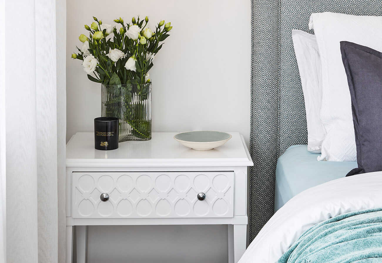 A white bedside table with a vase, candle and ceramic plate on it. 