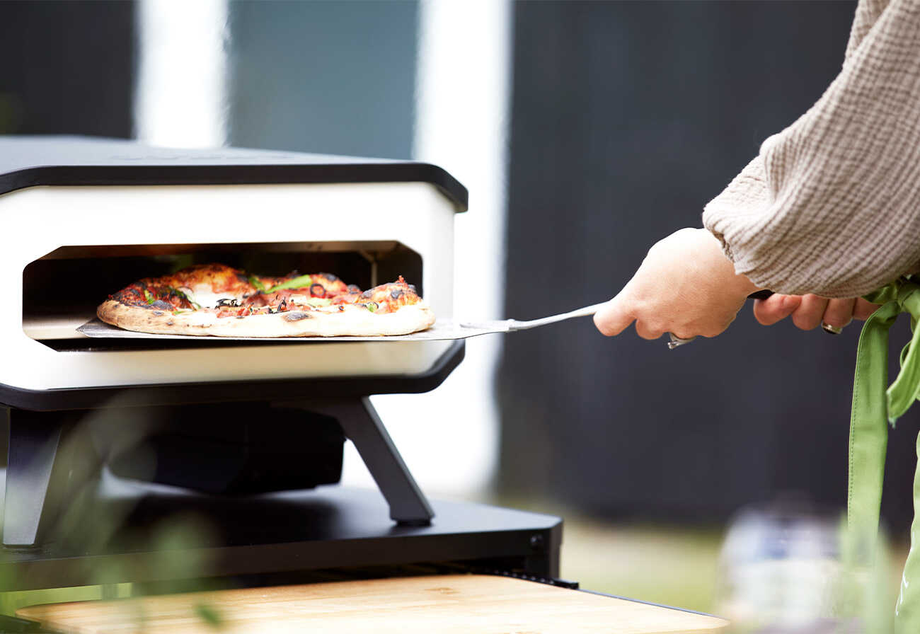 Person lifts a homemade pizza from the Cozze Electrical Pizza Oven.