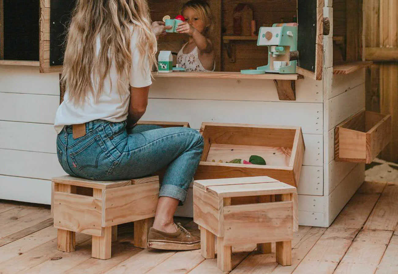 Woman sits on stools outside a kids' cubby house.