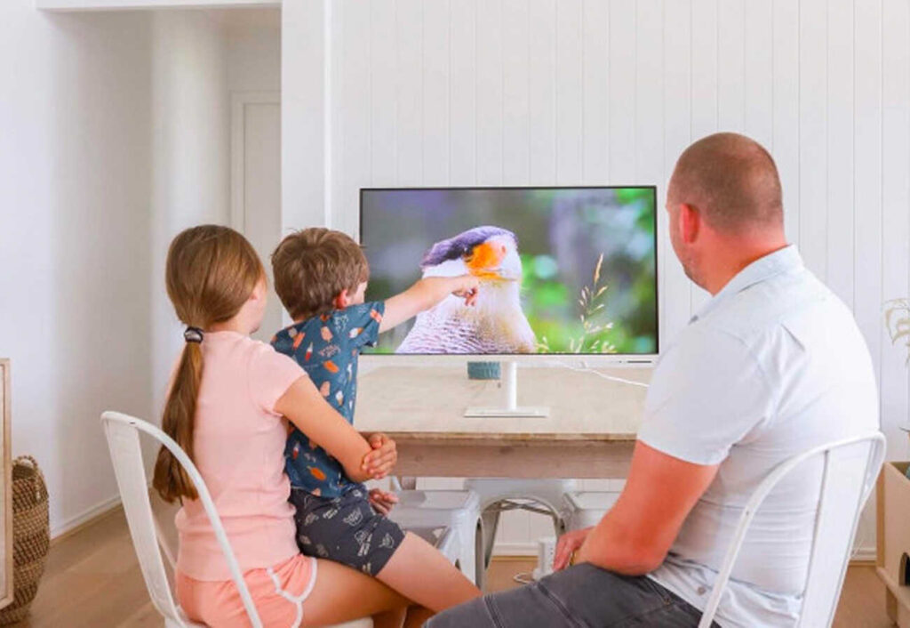 Two children and an adult man watch a bird on a large monitor.