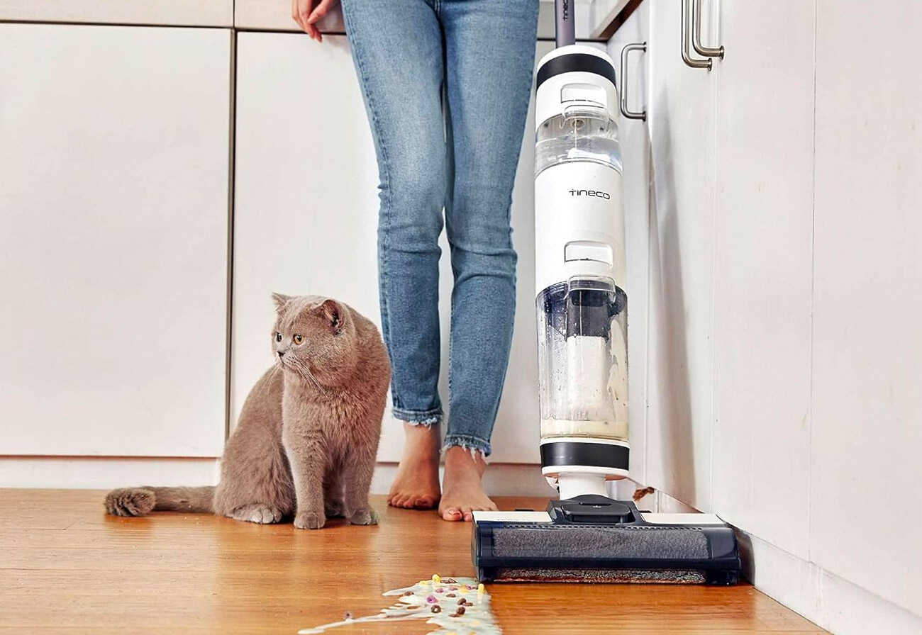 A cat sitting next to a lady as she vacuums a wet spill.