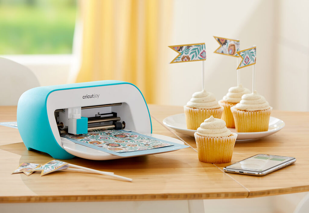 Cricut Joy machine in use on a table next to a plate of cupcakes.