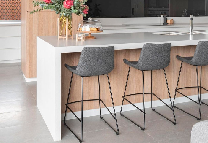 Modern kitchen with grey stone bench, grey stools and timber cabinets.