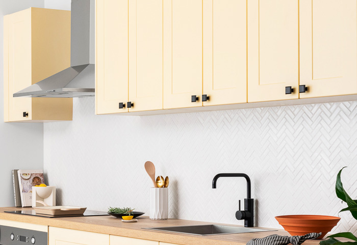 Pale yellow kitchen cabinets with black square-shaped knob handles and a black tap.