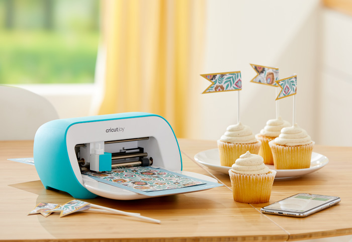 Cricut Joy on a table next to a plate of cupcakes.