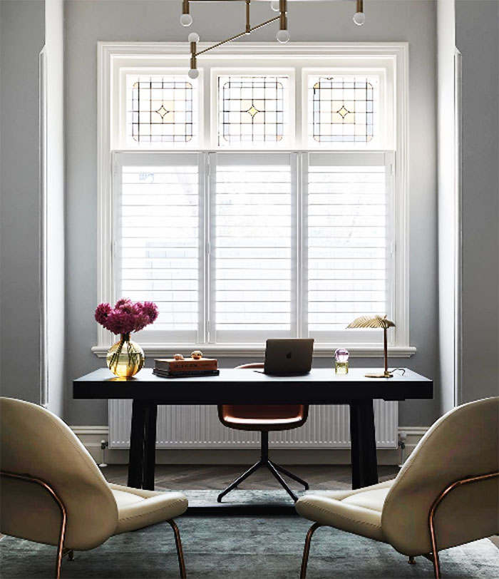 A home office with thick, dark wood desk, beige armchairs and a sage green rug. 