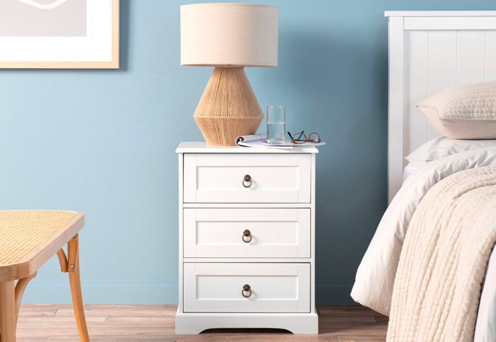 Classic white bedside table with a lamp, magazine and glass of water on top of it.