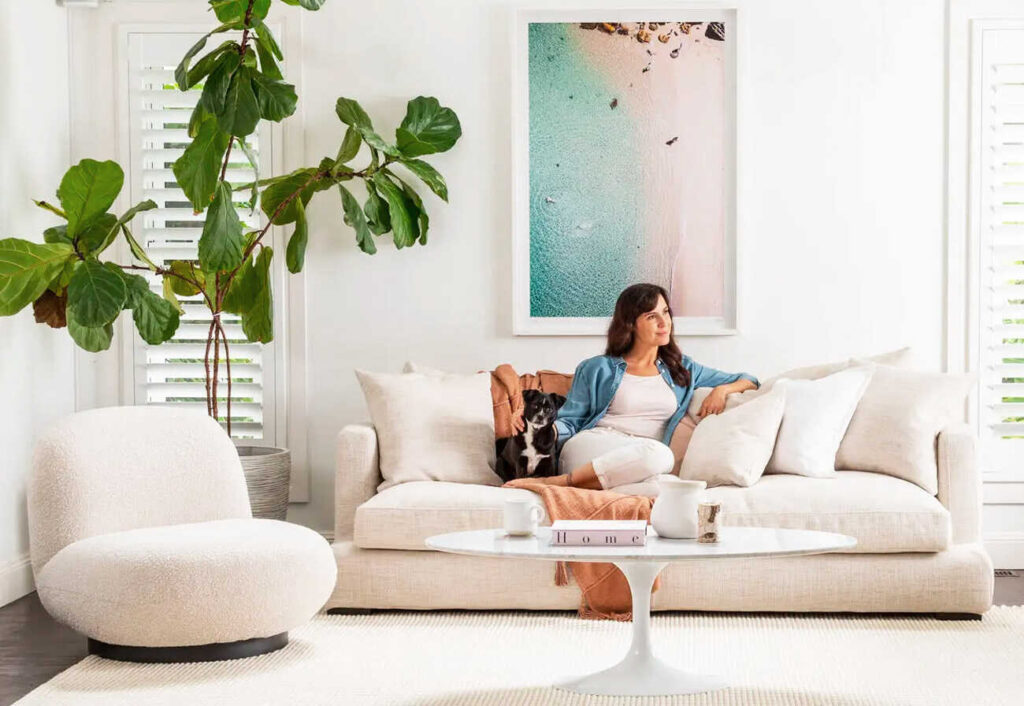 A woman and her dog sit on a natural-coloured sofa next to an indoor plant.