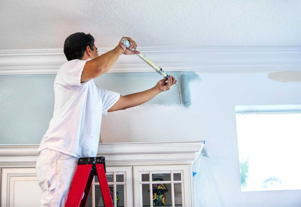 Main on a ladder painting an internal wall blue with a roller.