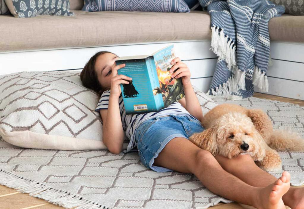 Child using a floor cushion as a pillow while she reads a book on a rug. 