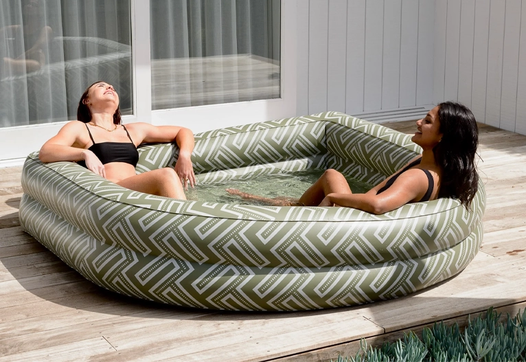 Two women lounging in a paddling pool on a deck.