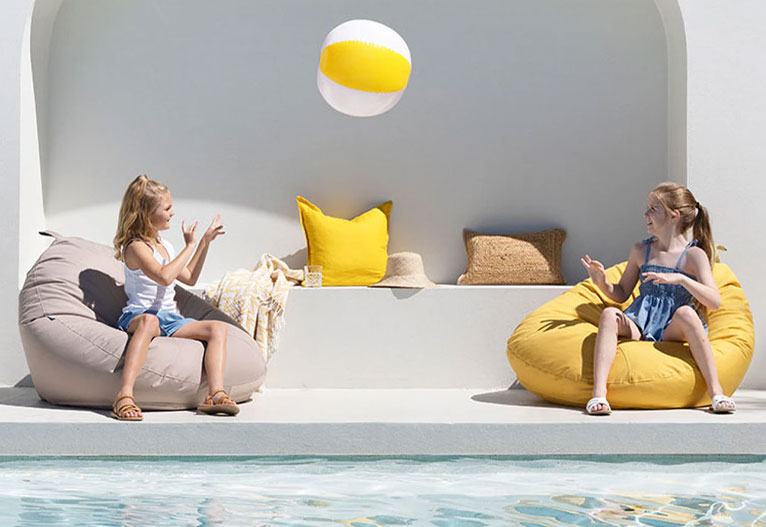 Children on bean bags play with a ball next to a pool.