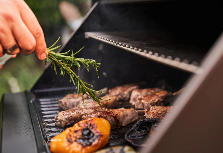 Person brushing meat and vegetables with a sprig of herbs on a BBQ plate.